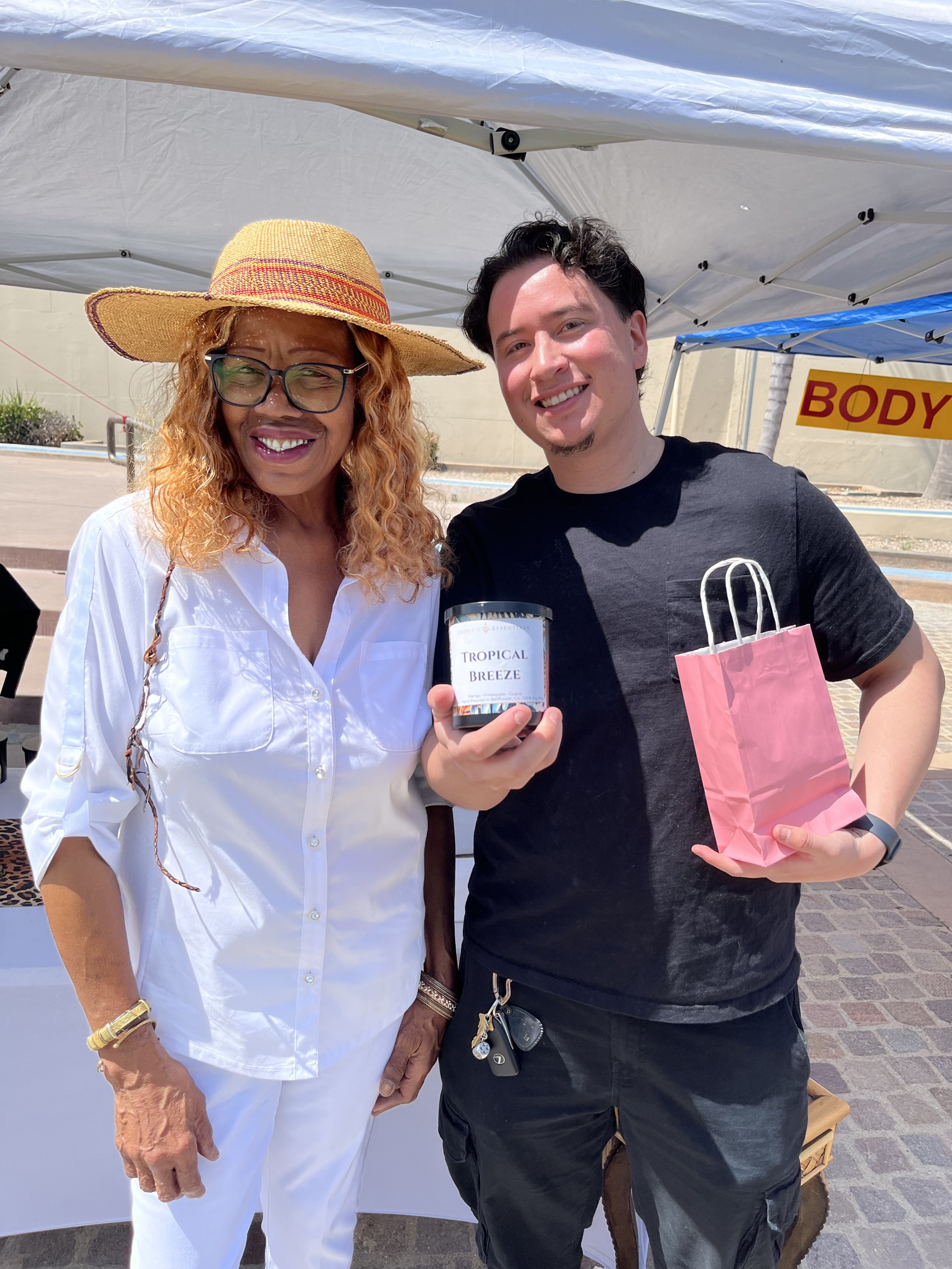 A young man holding a Tropical Breeze candle next to Debbie under the market tent