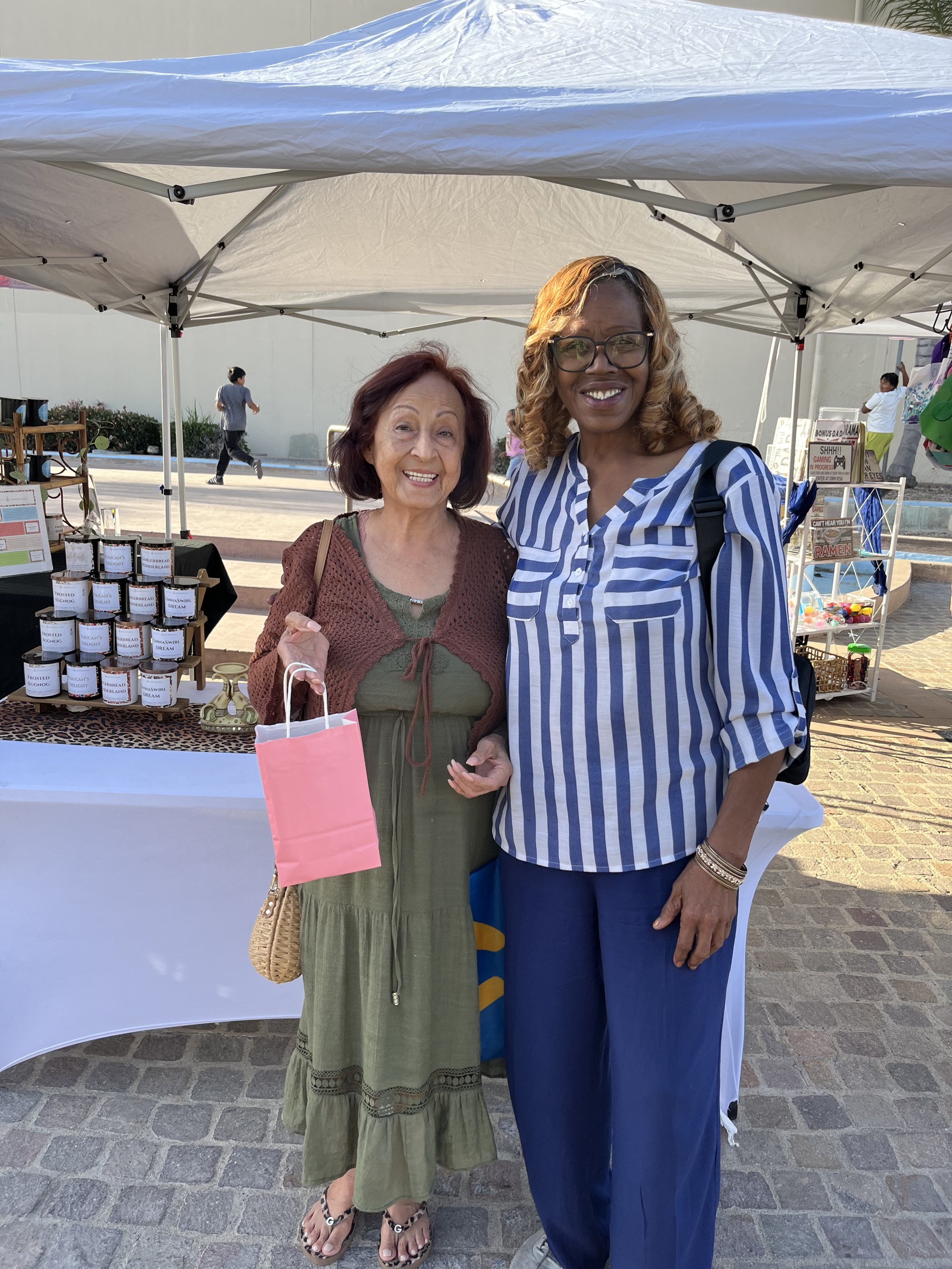 An older customer holding a pink Debbie's Essentials gift bag beside Deborah at the market
