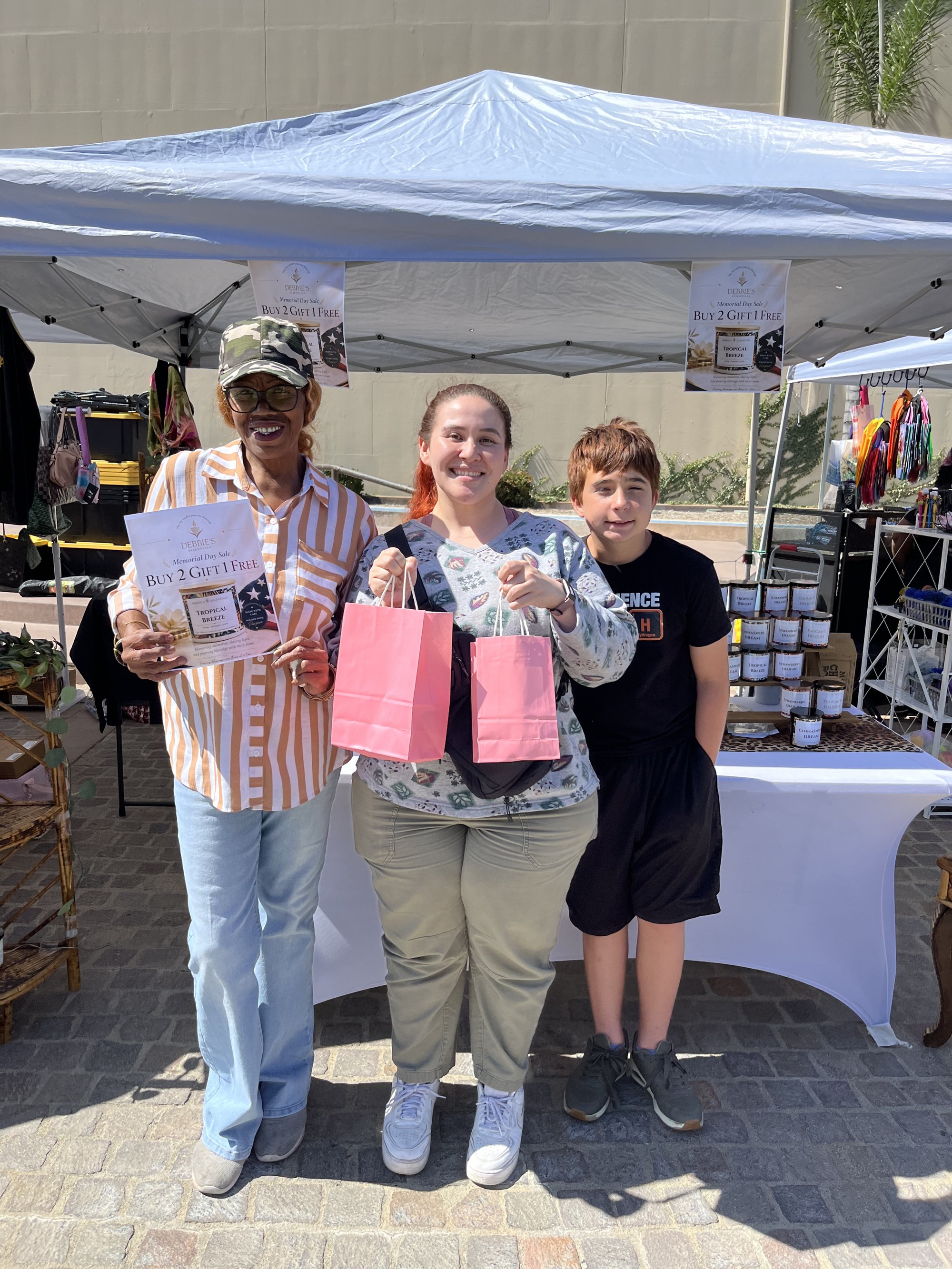 A mother, her son, and Deborah smiling with pink gift bags at the Memorial Day sale