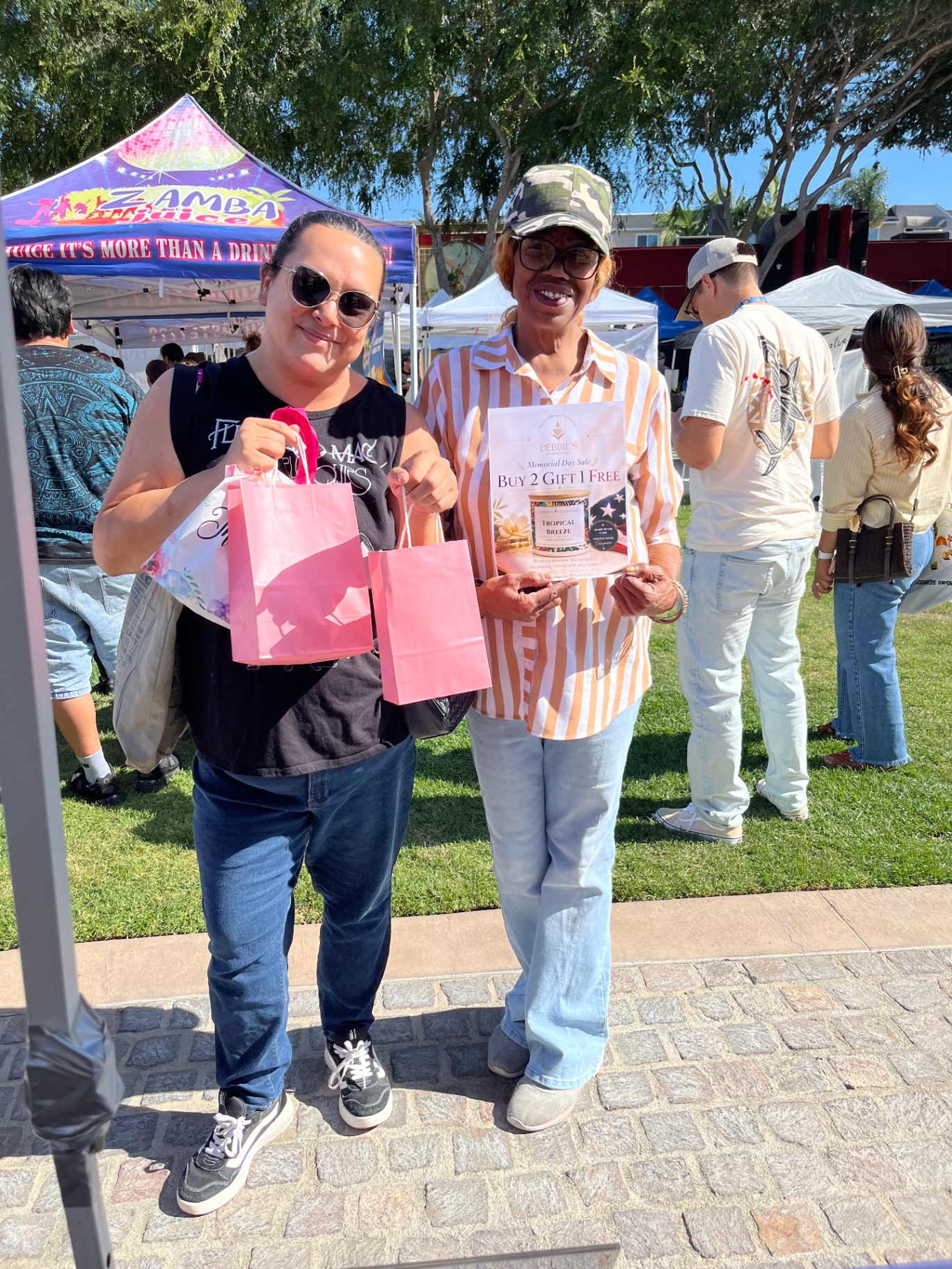 A customer in sunglasses holding two pink gift bags beside Deborah with a Tropical Breeze flyer at the outdoor market